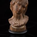 A wooden portrait bust sculpture set against a plain black background, mounted on a round black pedestal. The sculpture is constructed from numerous thin, vertical wooden slats arranged radially around a central axis, bound together near the top and base with thin light-colored cord. The carved edges of the slats collectively form the detailed contours of a person's head, face, and shoulder area.
