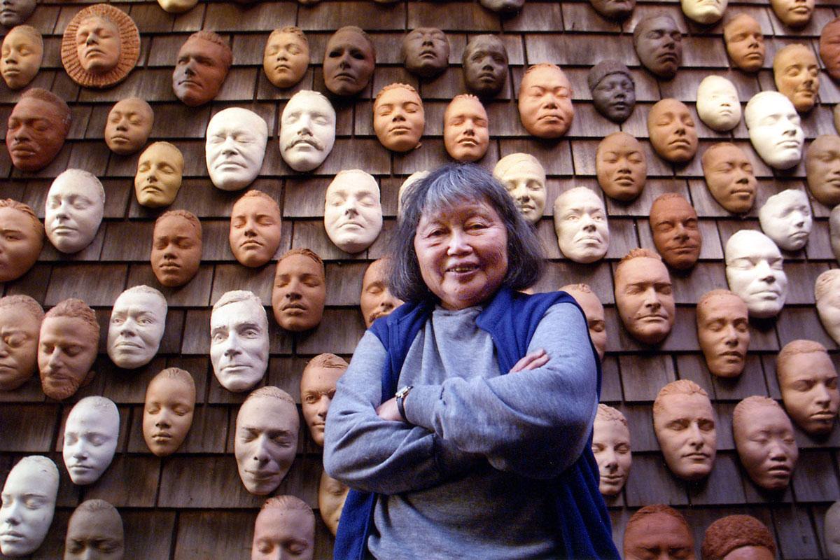 An image of artist Ruth Asawa infront of her Wall of Masks